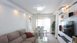Een moderne woonkamer met een beige bank, een glazen salontafel en een flatscreen televisie aan de muur.