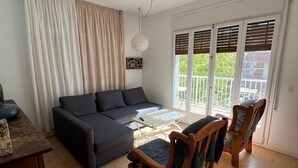 Ein Wohnzimmer mit einem dunkelgrauen Sofa, einem Holztisch und zwei Holzstühlen. Eine Glastür öffnet sich zum Balkon mit Blick auf Grünflächen.