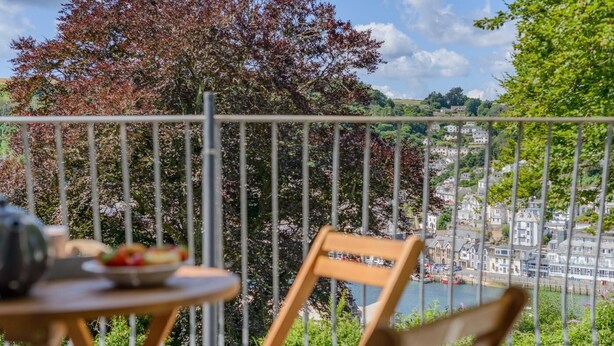 A balcony with a table and chair, overlooking a river and a town with buildings and trees. - 2 Bed in Looe (oc-g32529) (Looe)