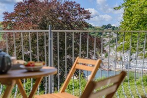 Un balcone con tavolo e sedia, affacciato su un fiume e una cittadina con edifici e alberi.
