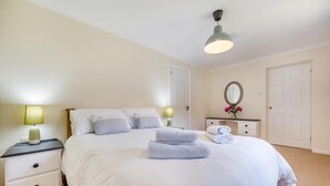 A neatly made bed with white linens and folded towels, a bedside lamp, a dresser with a mirror, and a door in the background. - 2 Bed in Looe (oc-g32529) (Looe)