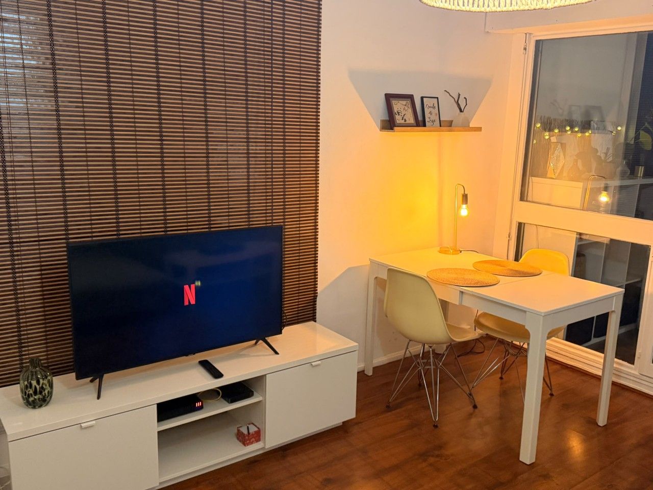 A modern living room with a flat-screen TV, a dining table, chairs, and a window with blinds.