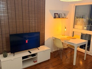 A modern living room with a flat-screen TV, a dining table, chairs, and a window with blinds.