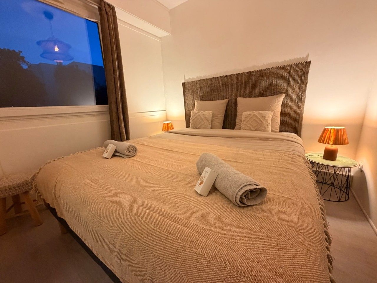 A hotel room with a large bed, two bedside lamps, a window, and a small table.