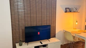 Een moderne woonkamer met een flatscreen televisie, een witte entertainmentkast, een eethoek met een tafel en stoelen, en een wandplank met decoratieve objecten.