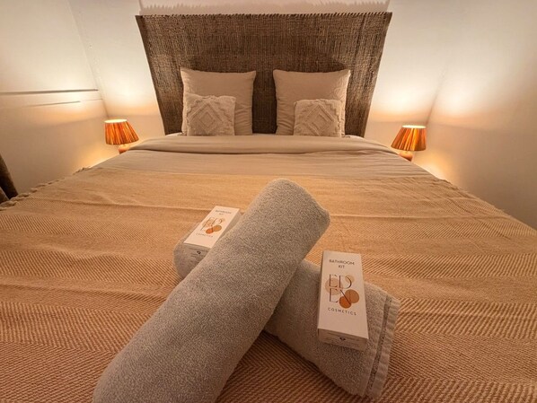 Un lit bien fait, avec une couvre-lit beige et des oreillers, deux serviettes enroulées et une lampe de chevet de chaque côté.