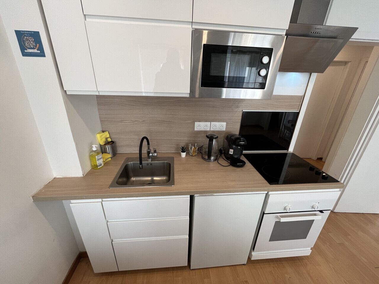 A compact kitchen with a stainless steel microwave, a built-in oven, a sink, and a coffee maker.