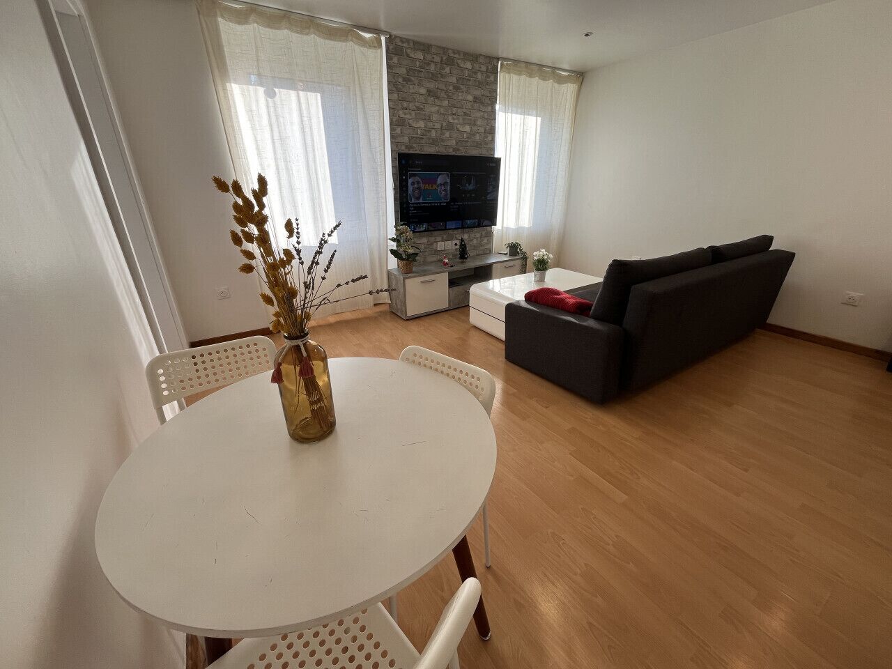 Ein Wohnzimmer mit einem weißen runden Tisch, einer Couch, einem Fernseher und einem Fenster mit Vorhängen.