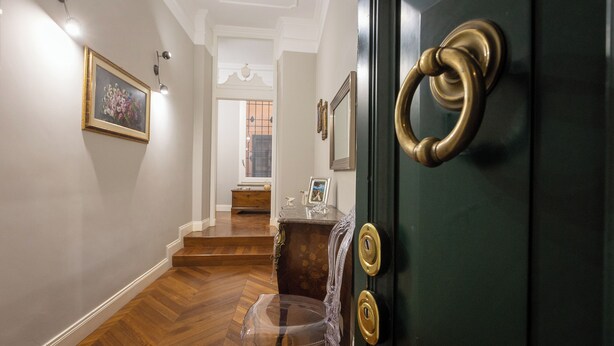 A hallway with a wooden floor, a green door with a brass ring handle, a framed picture on the wall, and a small table with a picture frame and a vase. - Rental in Rome Gregoriana Apartment (Rome)