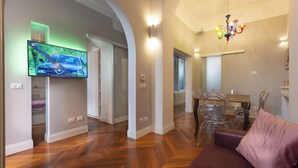 A modern living room with a flat-screen TV, wooden flooring, a dining area with clear chairs, and a chandelier. - Rental in Rome Gregoriana Apartment (Rome)