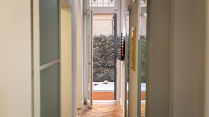 A hallway with a wooden floor, a door with a security camera, and a view of a garden with a stone wall and a bench. - Rental in Rome Gregoriana Apartment (Rome)