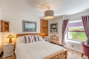 A bedroom with a bed, bedside tables, a lamp, a window with curtains, a bookshelf, and a framed picture on the wall.