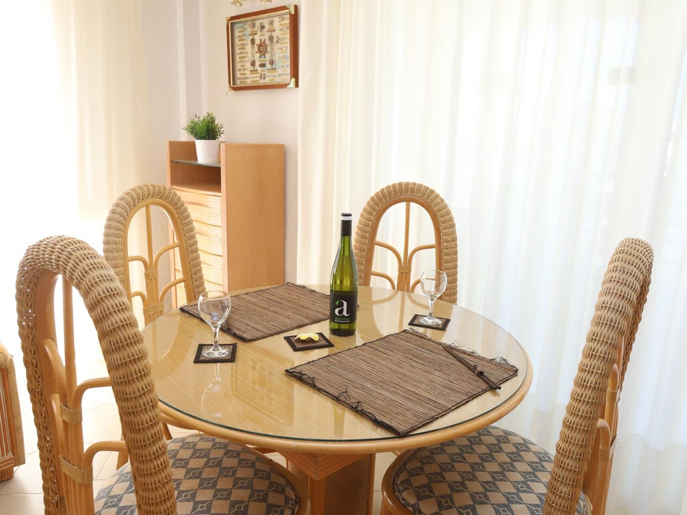 A round glass table with four wicker chairs, each with a patterned cushion. A bottle of wine and two wine glasses are on the table.