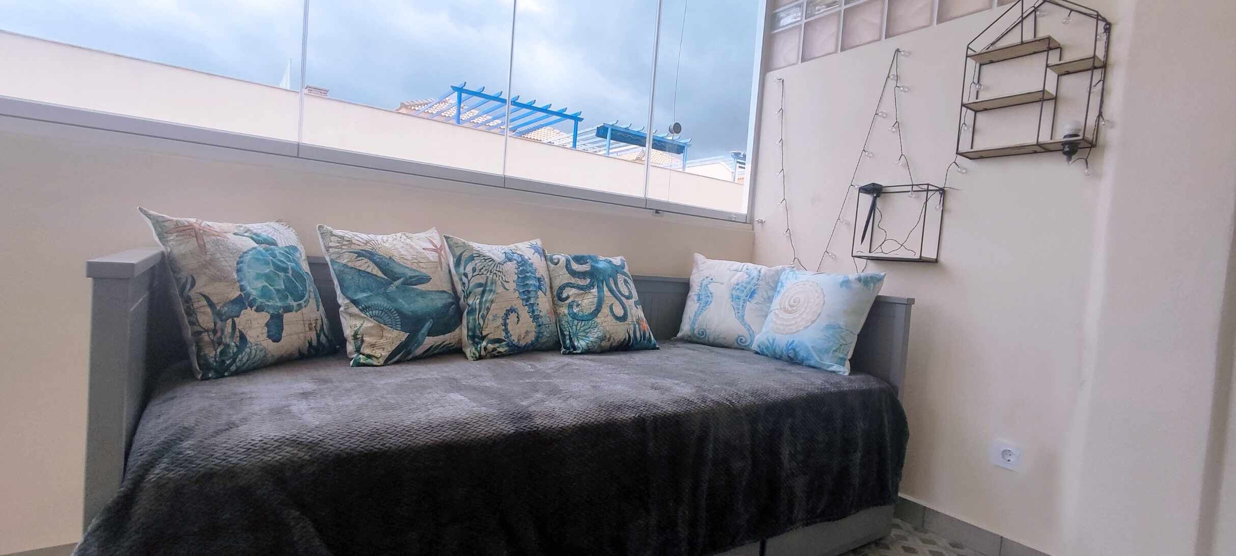 Un lit recouvert d’une couverture sombre et agrémenté de quatre coussins décoratifs, une fenêtre donnant sur une structure bleue et une étagère fixée au mur sur laquelle est posée une petite plante.