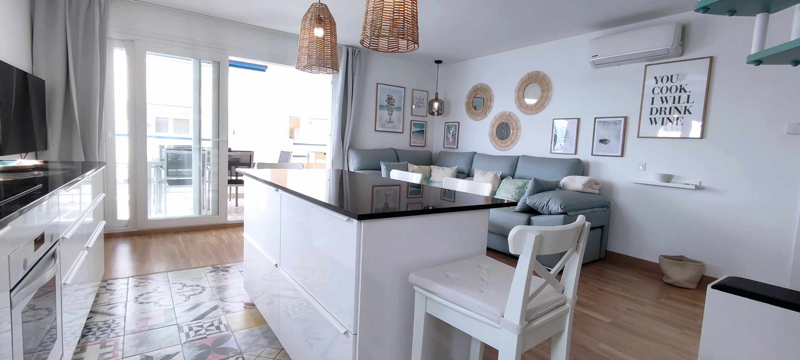 A modern kitchen with a dining area, featuring a white countertop, a dining table, and a sofa. There are decorative wall mirrors and a framed quote on the wall.
