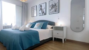 A neatly made bed with blue bedding, a bedside table with a lamp, and a large mirror on the wall. - Albaida Gardens (El Morche)