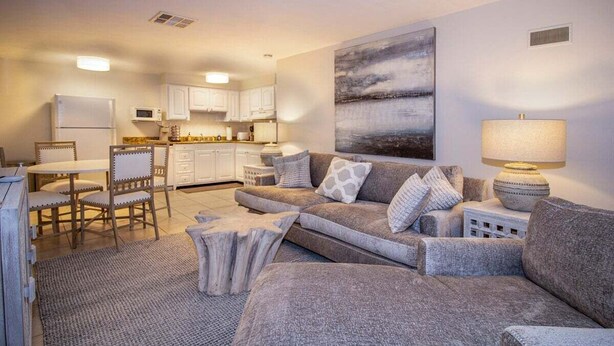 Una sala moderna con un sofá gris, una mesa de comedor con sillas, una zona de cocina y un cuadro abstracto grande en la pared.