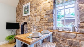 A stone-walled room with a wooden table set for dining, a television mounted on the wall, and a window with a view of greenery outside. - 1 Bed in Drumnadrochit (oc-t32404) (Drumnadrochit)