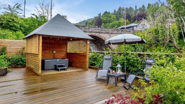A wooden deck with a hot tub and lounge chairs, surrounded by lush greenery and a stone bridge in the background. - 1 Bed in Drumnadrochit (oc-t32404) (Drumnadrochit)