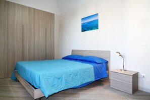 Ein Schlafzimmer mit einem Bett, einem Nachttisch, einer Schublade, einer Wandlampe und einem Bild vom Meer.