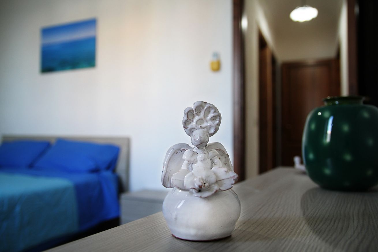 Une statuette d’ange en céramique blanche est posée sur une table en bois, avec un lit bleu et un vase vert en arrière-plan.