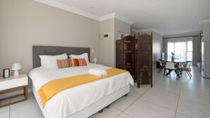 A hotel room with a large bed, a bedside table with a lamp, a wooden partition, and a dining area with chairs and a table.