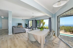 A modern living room with a dining table, grey sofa, and large windows overlooking a pool and ocean. - Spacious Family Camps Bay Escape |  Spectacular Views | Pool, Garden (Cape Town)