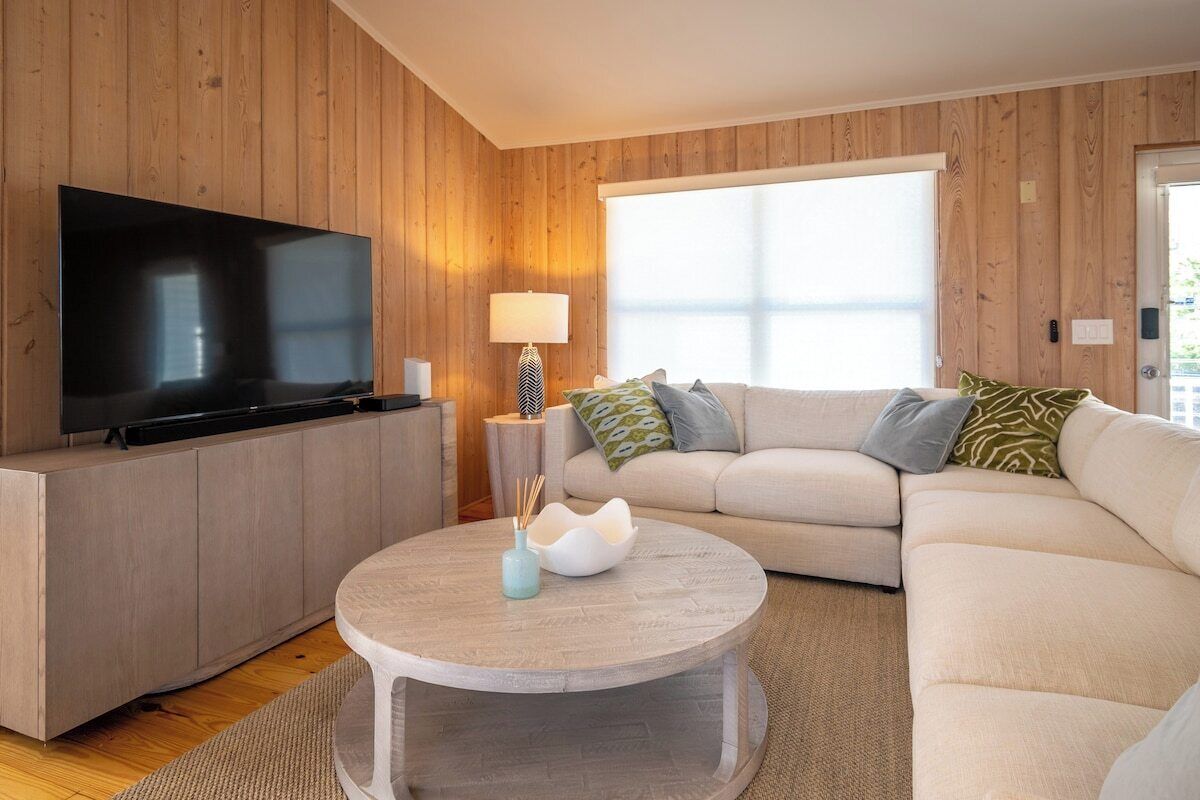 A modern living room with a flat-screen TV, a round coffee table with decorative items, a beige sofa with patterned cushions, and a wooden paneled wall.