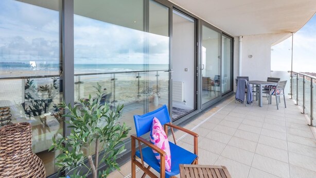Un balcon avec vue sur la plage, une table avec des chaises et un fauteuil avec un coussin.