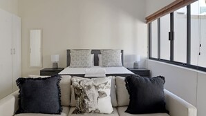 A bedroom with a bed, two bedside tables, a window, and a sofa with pillows. - Modern Sea Point Oasis | Garden, Pool, BBQ & Gym (Cape Town)