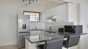 A modern kitchen with white cabinets, a stainless steel refrigerator, a microwave, and a coffee maker. There is a window with blinds and a door to the left. - Modern Sea Point Oasis | Garden, Pool, BBQ & Gym (Cape Town)