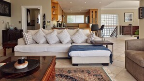 Ein geräumiges Wohnzimmer mit einer Couch, Hocker und Couchtisch. Im Hintergrund ist eine Küche mit Holzschränken und einem Essbereich zu sehen.