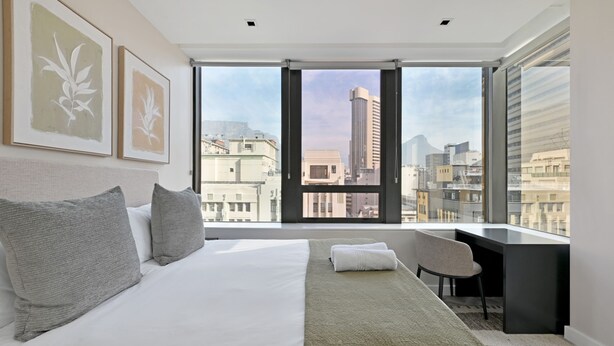 Una habitación de hotel moderna con una cama grande, un escritorio y vistas a la ciudad a través de ventanales de pared a techo.