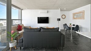 Ein modernes Wohnzimmer mit einer schwarzen Couch, einem Essbereich mit weißem Tisch und Stühlen sowie einem großen Fenster mit Blick auf die Stadt.