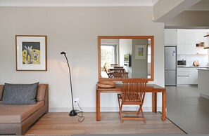 A modern living room with a sofa, a dining table with chairs, a floor lamp, and a large mirror. A kitchen is visible in the background. - Cozy 2-Bed Apartment Below Table Mountain (Cape Town)