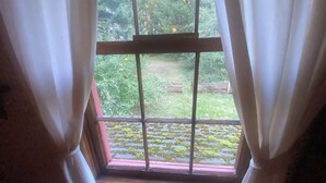 A window with white curtains, a view of a garden with grass and trees, and a wooden floor.