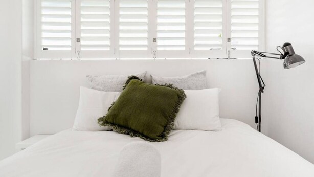 Cama bem arrumada com roupa de cama branca e travesseiro verde, uma luminária de cabeceira e persianas brancas na janela.