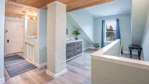 Une pièce avec un plafond en pente, un escalier, une porte blanche, un banc en bois et une plante.