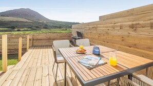 Une terrasse extérieure en bois, avec une table dressée pour le petit-déjeuner, un barbecue et une vue sur une montagne.