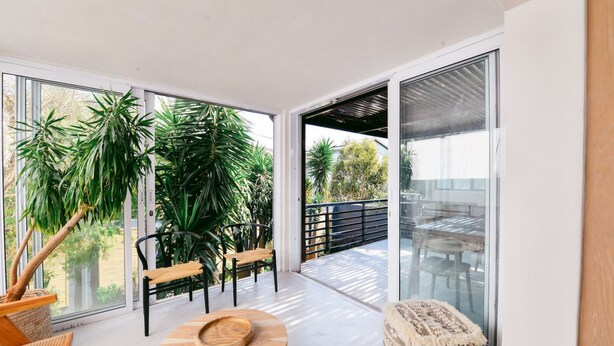 Een modern interieur met een ronde houten tafel, twee stoelen en een geweven poef. Grote ramen en een balkon met glazen hekwerk zijn zichtbaar.