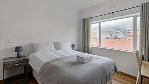 Una habitación de hotel moderna con una cama grande, una mesita de noche con lámpara, una silla y una ventana con vistas a la ciudad.