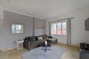 Un soggiorno moderno con un divano grigio, un tavolino bianco, un tavolino rotondo con un vaso di fiori, un'ampia finestra con tende e un televisore a schermo piatto.