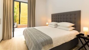 Hotelkamer met een groot bed, een grijs hoofdeinde, twee bedlampen en uitzicht op een gele muur en een balkon.