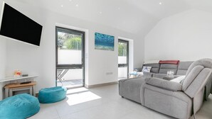 Ein modernes Wohnzimmer mit einem grauen Sofa, türkisfarbenen Sitzsäcken, einem Flachbildfernseher und einem großen Fenster mit Blick ins Freie.