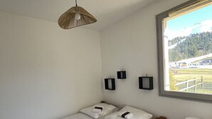 Een enkel bed met witte beddengoed, een hangende geweven lamp en drie kleine, zwartgekleurde wandlampen. Een raam toont uitzicht op een groen landschap met een hek en gebouwen.