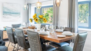 Comedor con mesa de madera dispuesta para seis personas, rodeada de sillas negras. Tres lámparas colgantes sobre la mesa, y un jarrón con girasoles sobre la misma.