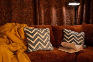 A cozy room with a patterned wallpaper, a brown sofa, two patterned pillows, a yellow blanket, and an open book. - 4 Bed in Woolfardisworthy (oc-s32421) (Higher Clovelly)
