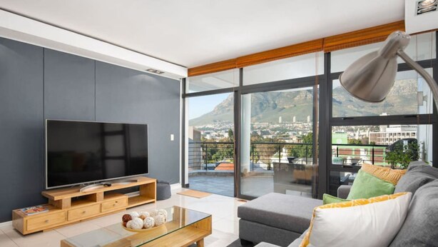 A modern living room with a large flat-screen TV, a glass coffee table, and a grey sofa with colorful cushions. - 8th Floor- Table Mountain Views|Central & Secure (Cape Town)