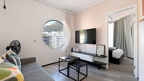 A modern hotel room with a round window, a flat-screen TV, a bed, a desk, and a sofa. - The Flamingo | One Bed | Sea Point Promenade (Cape Town)