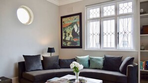 A living room with a dark grey sofa, a white coffee table with books and a vase of flowers, a large poster on the wall, and a window with white bars. - Spacious 3 Bedroom Home | Green Point (Cape Town)